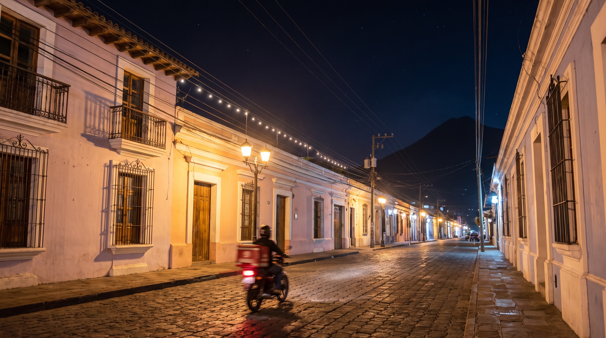 Calle de casas coloniales de noche con una moto de reparto y un volcán al fondo